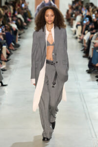 Model walks a runway in an oversized gray suit with a white coat draped, revealing a gray bralette and belt.