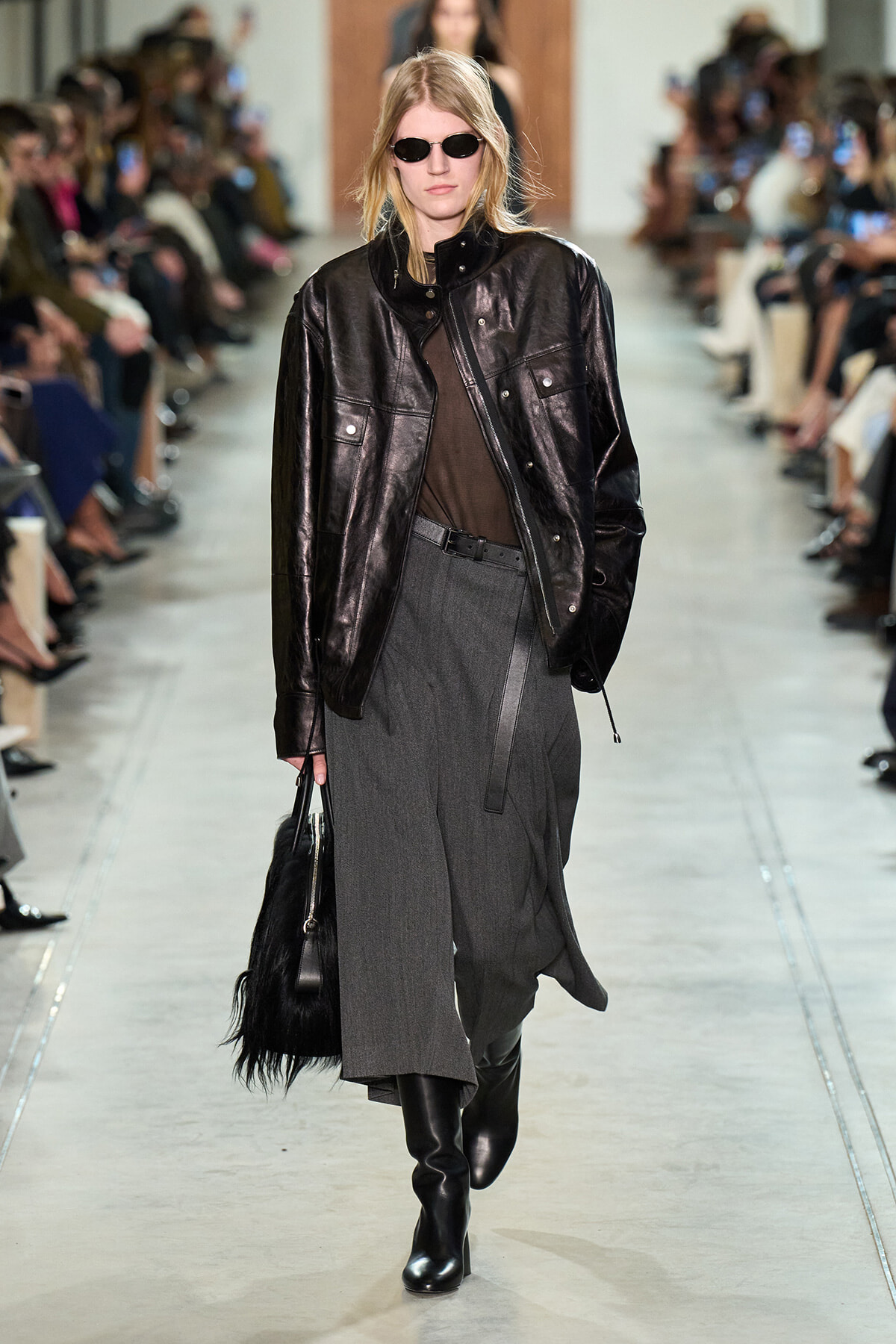 Model walking a runway in a shiny black leather jacket, brown sheer top, gray wide-leg skirt, black boots, and sunglasses, carrying a black bag with fringe fielding audience in background.