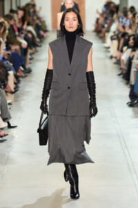 Model walks a runway in a gray sleeveless blazer over a black turtleneck, matching gray skirt, black gloves, and tall black boots, carrying a black handbag.