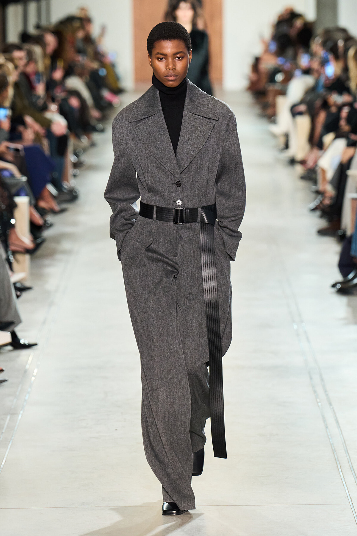 Female model walks a runway in a charcoal gray tailored suit with wide lapels, black turtleneck, and a wide belt.