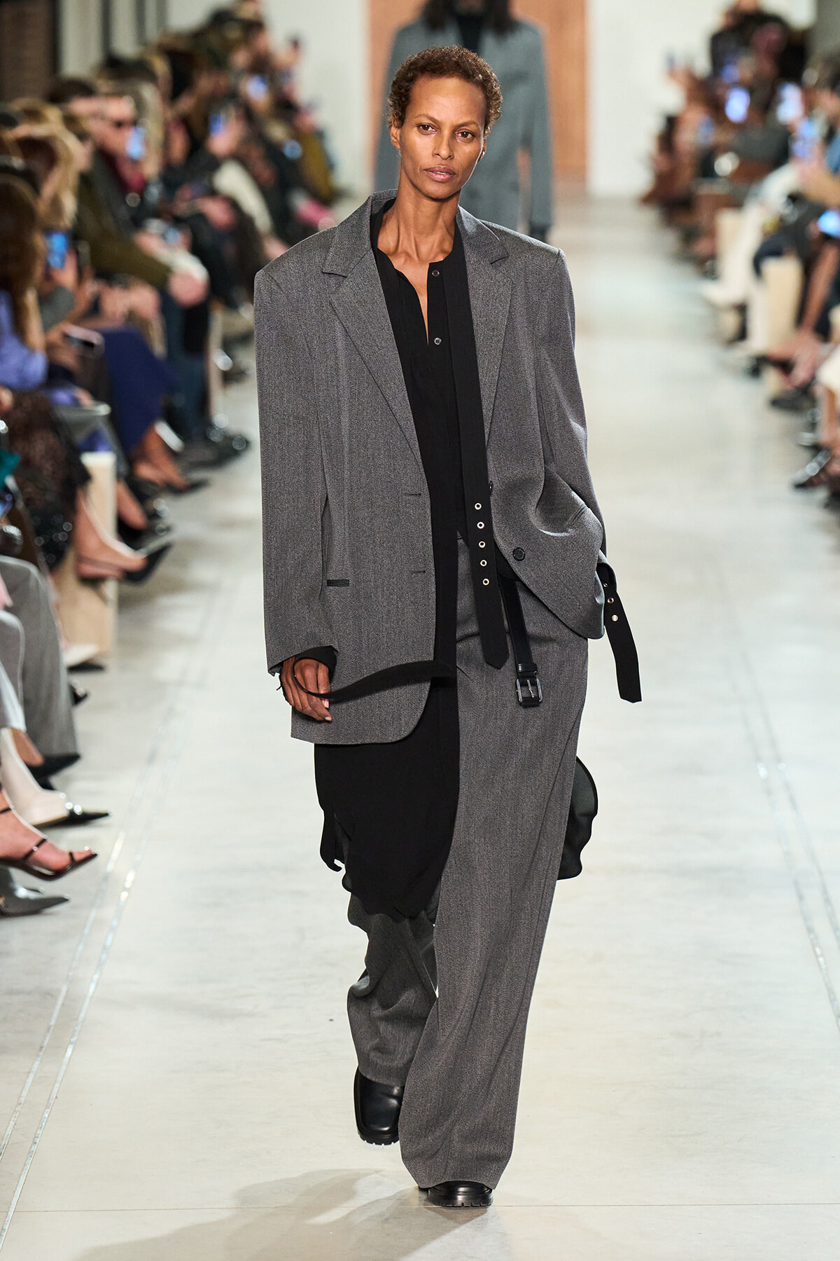 Model walking a fashion runway in an oversized gray suit over a black blouse, audience on both sides watching.