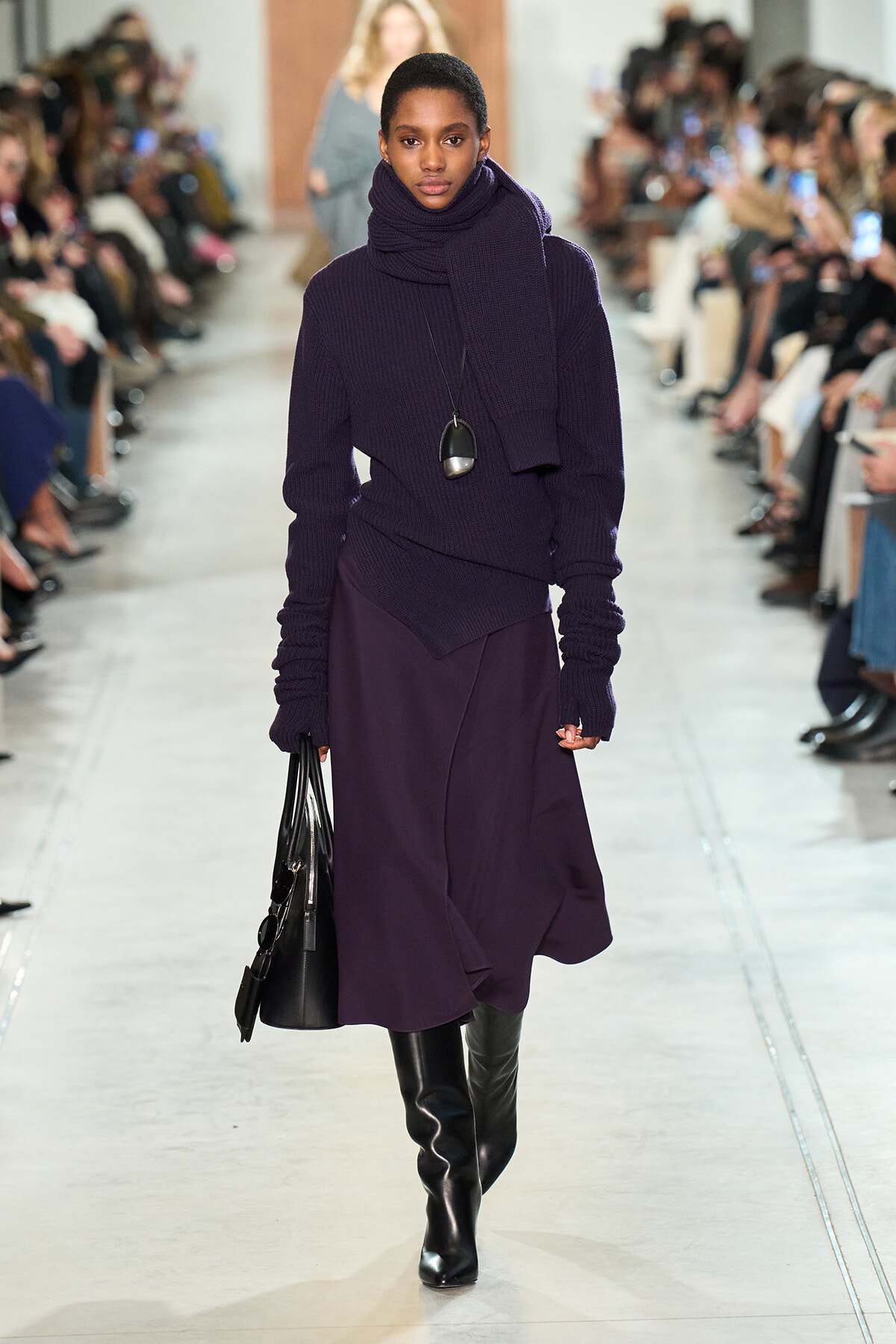 Model walking a runway in an all-purple outfit: chunky knit sweater with a large scarf, a layered midi skirt, and knee-high black boots, carrying a black handbag.