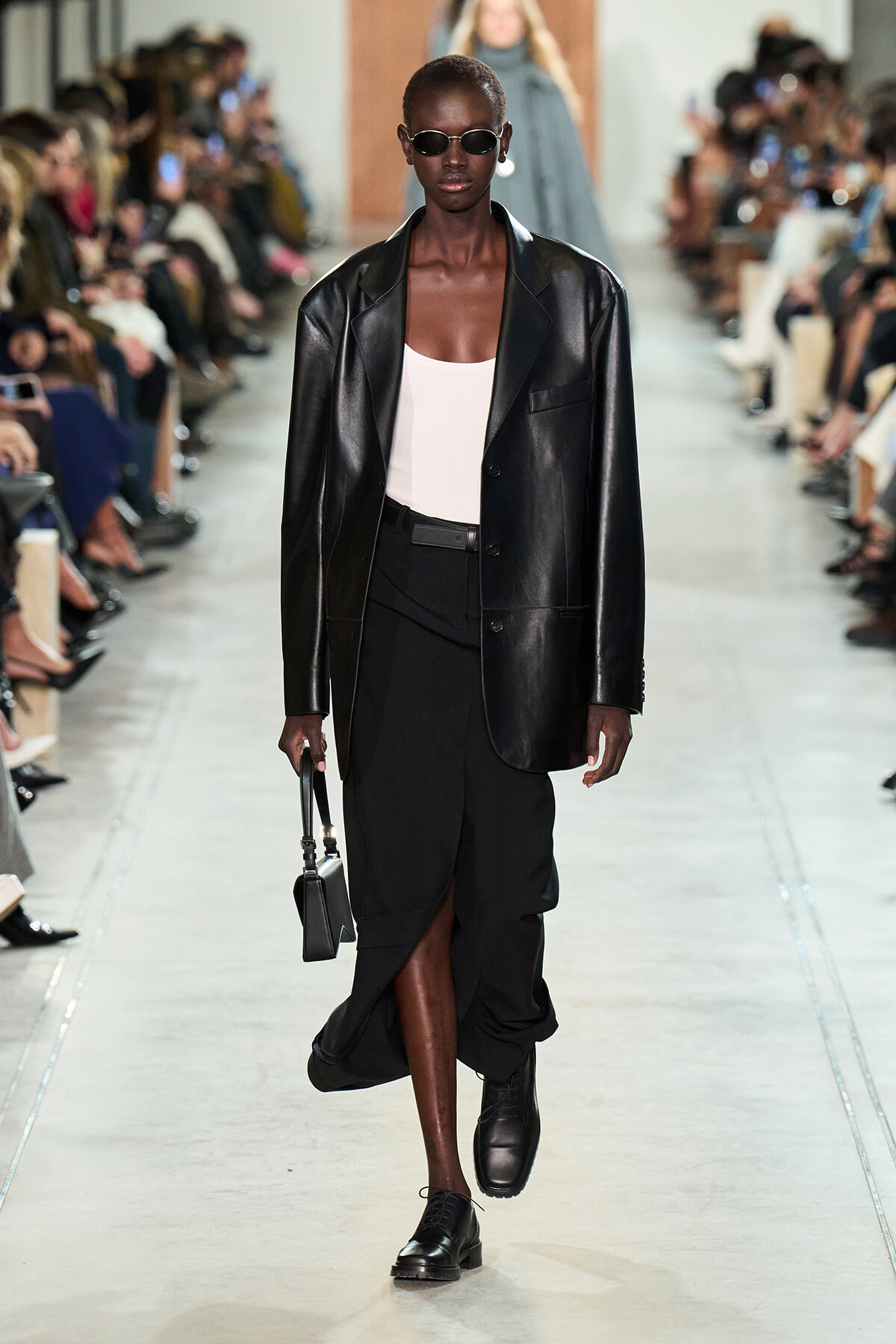 Model walks runway in an oversized black leather blazer, white top, black high-waisted skirt with a front slit, black shoes, sunglasses, and a small black handbag.