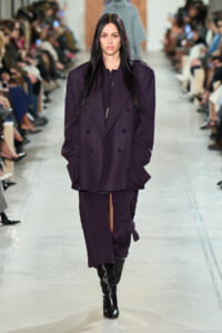 Model walking the runway in a dark purple oversized double-breasted blazer and matching pants with black knee-high boots.