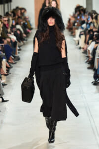 Model walking a runway in an all-black ensemble with a large black fur hood, knee-high boots, and a black structured bag.