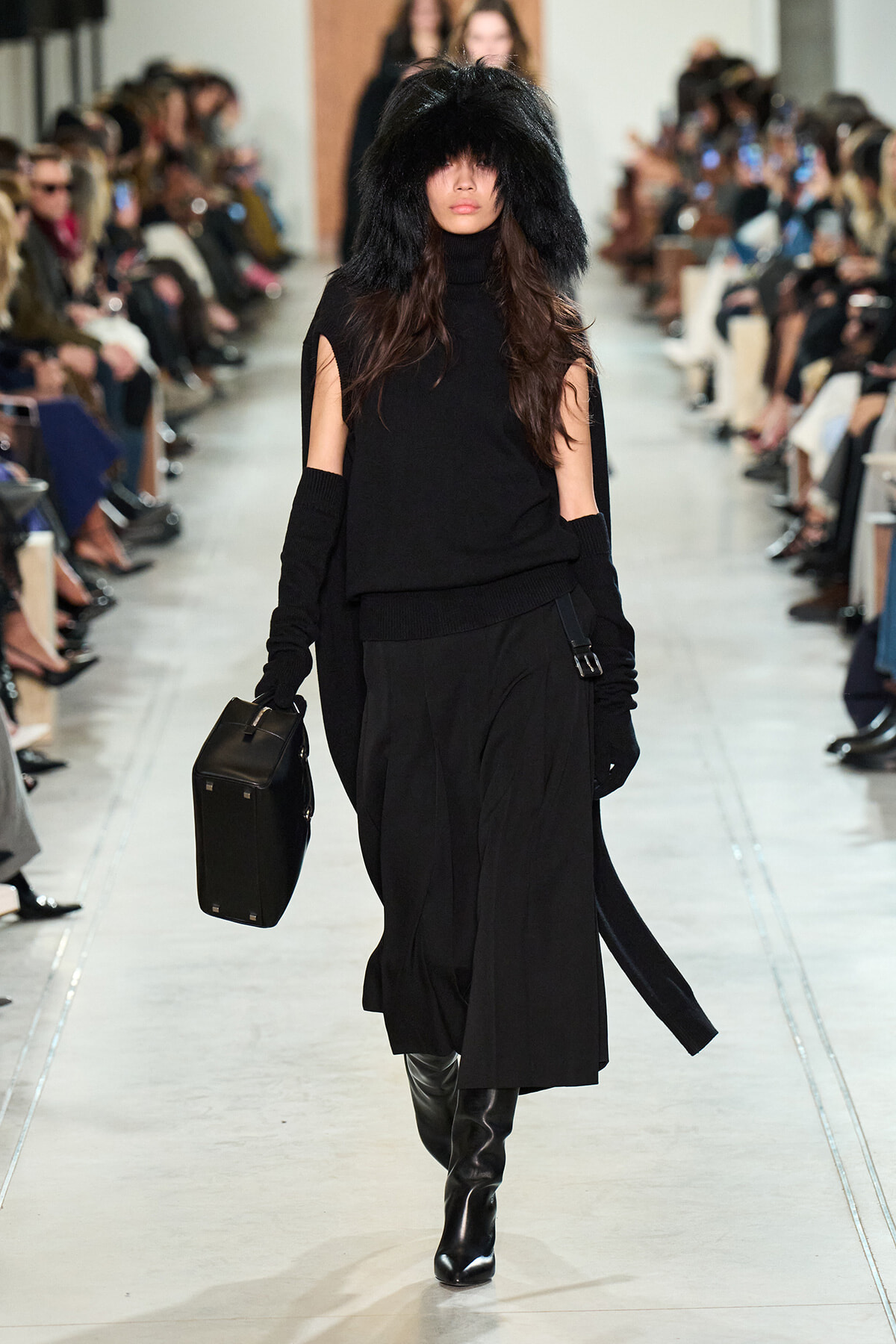Model walking a runway in an all-black ensemble with a large black fur hood, knee-high boots, and a black structured bag.