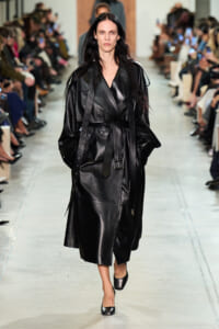 Model walking the runway in a black leather trench coat with a waist belt and wide sleeves, audience on both sides watching.