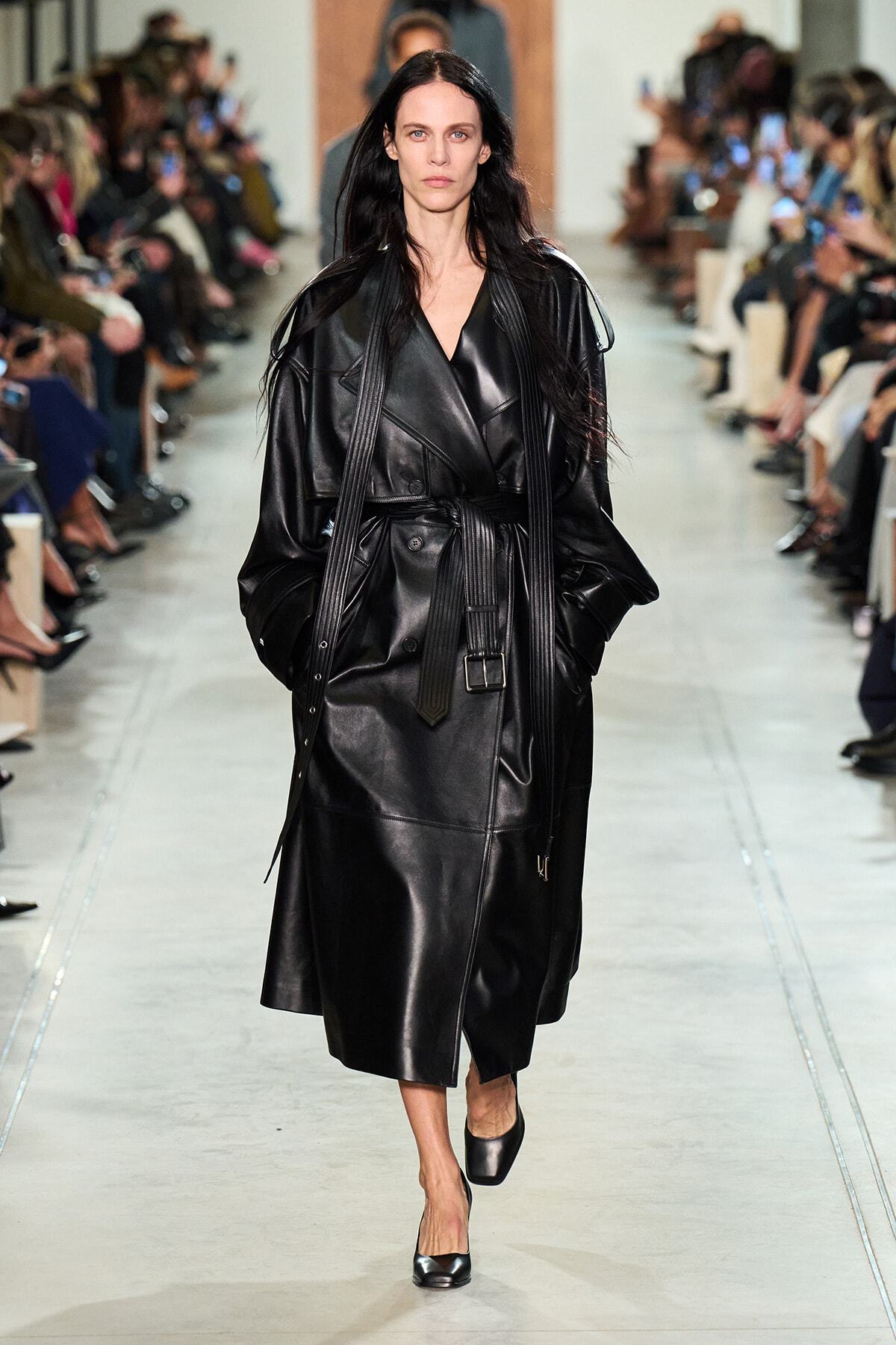 Model walking the runway in a black leather trench coat with a waist belt and wide sleeves, audience on both sides watching.