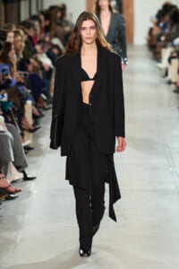 Female model walks a runway in a black tailored blazer over a black bralette and layered, asymmetrical skirt with black pants and shoes, holding a black clutch.