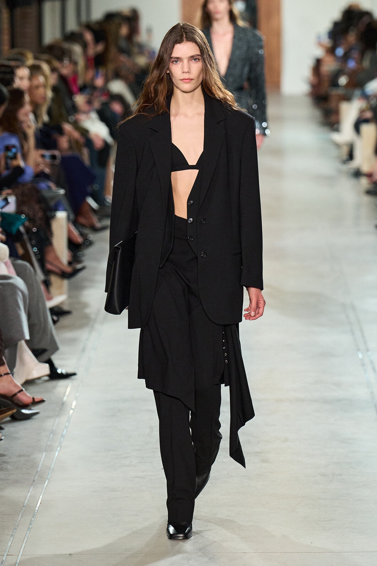 Female model walks a runway in a black tailored blazer over a black bralette and layered, asymmetrical skirt with black pants and shoes, holding a black clutch.