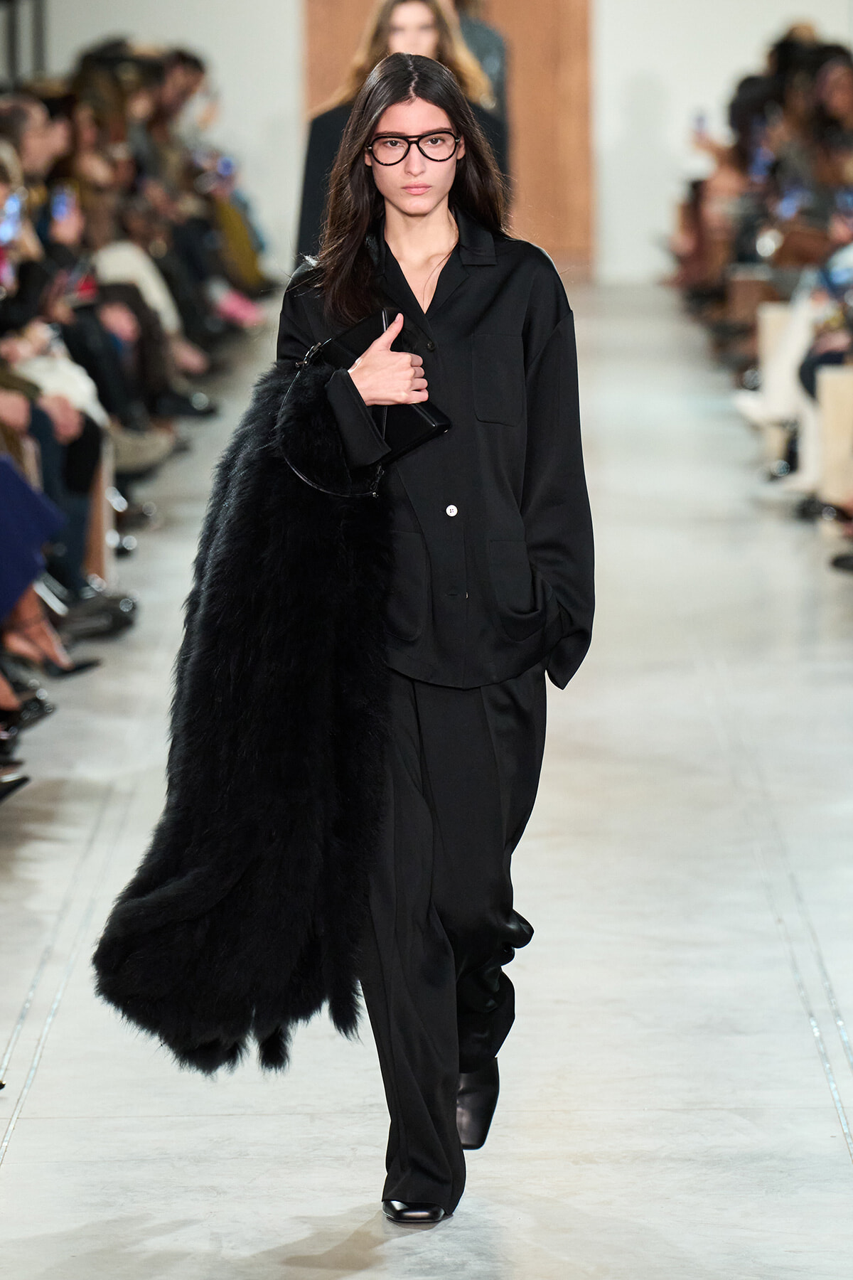 Model walks a runway in an all-black outfit—long coat, wide trousers, and a long black fur stole draped over one arm, holding a black clutch; audience line the runway in the background.