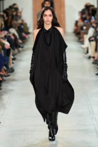 Model walks the runway in a loose, draped black dress with cape-like panels, paired with long black gloves and knee-high boots.