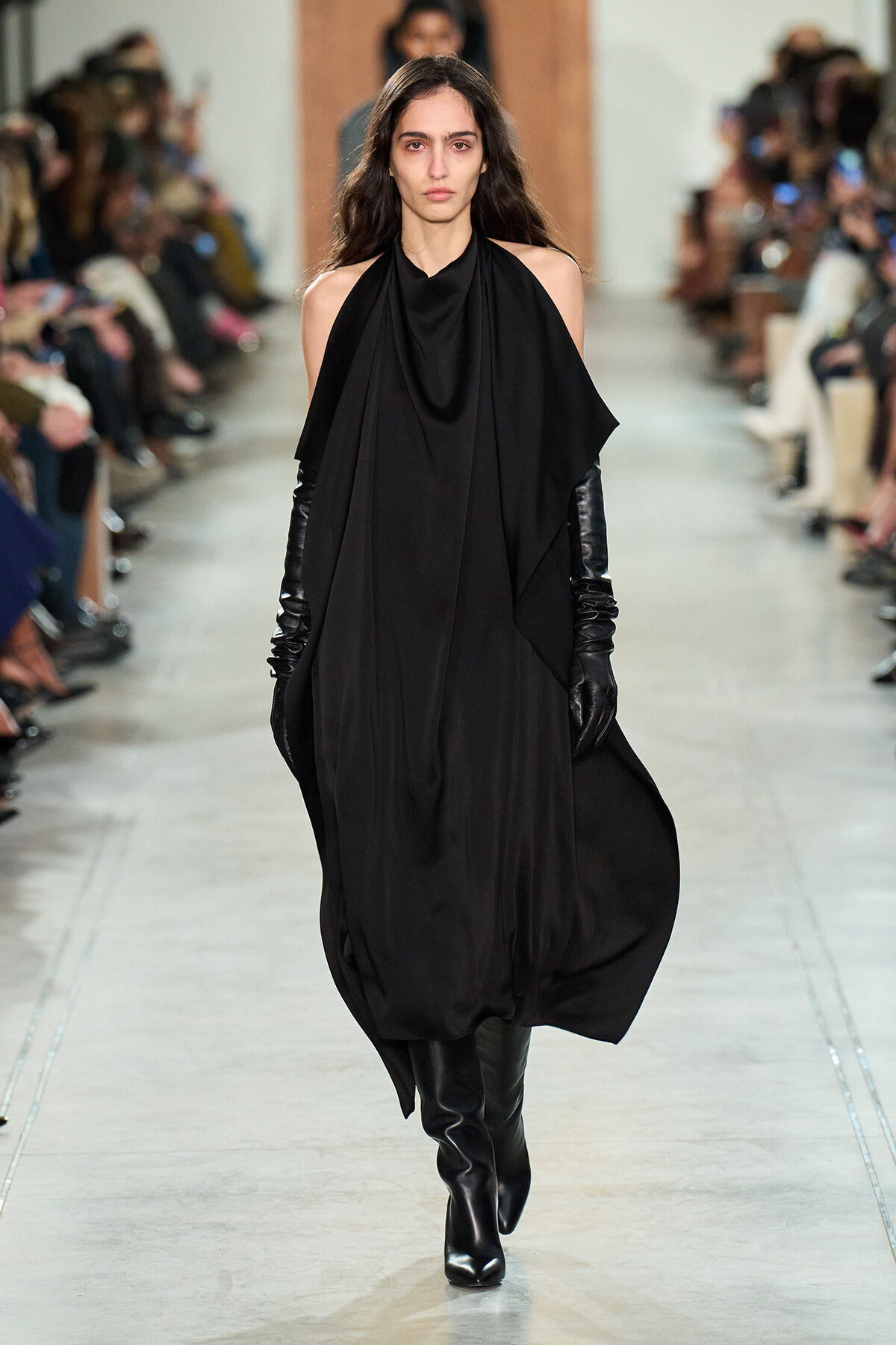 Model walks the runway in a loose, draped black dress with cape-like panels, paired with long black gloves and knee-high boots.