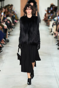 Model walking a fashion runway in a black fur-like coat, black skirt, gloves, and ankle boots, carrying a black bag.