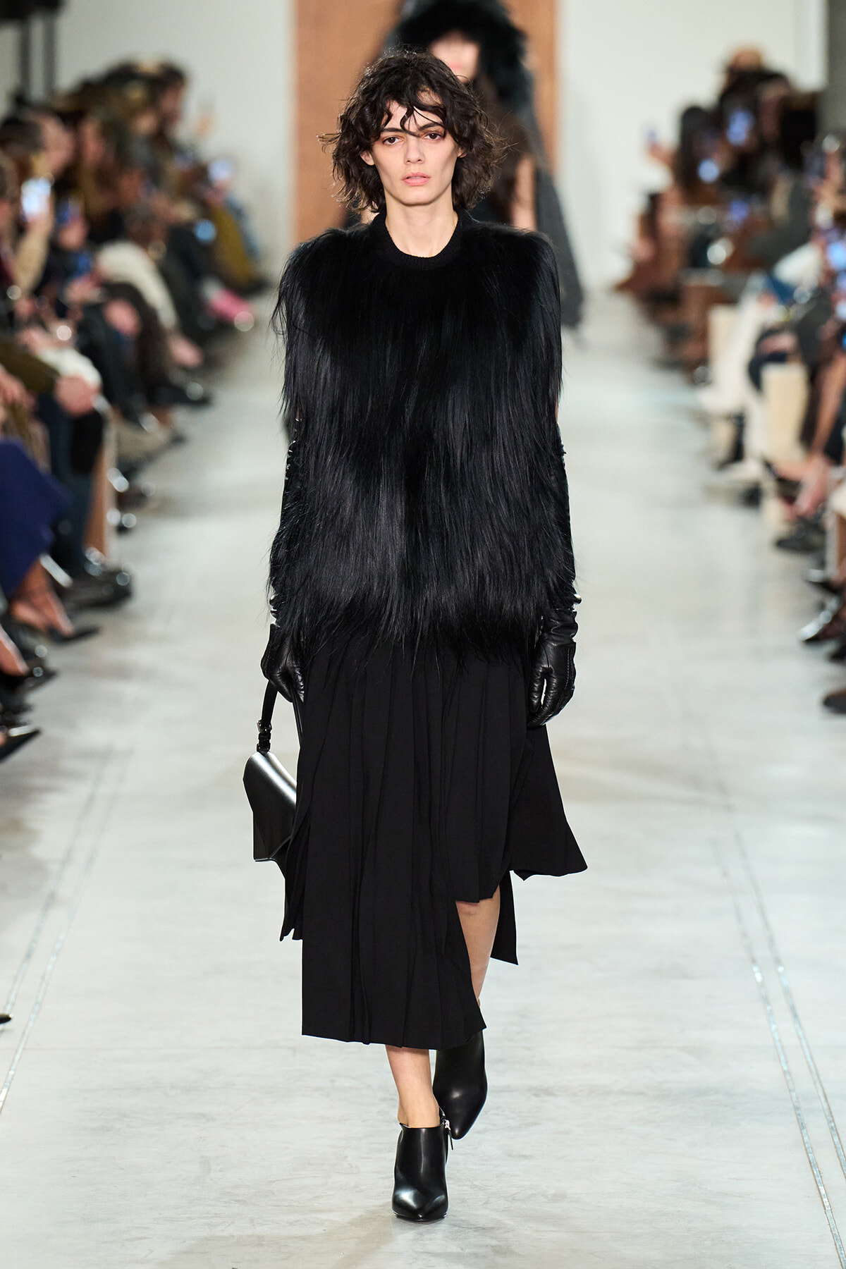 Model walking a fashion runway in a black fur-like coat, black skirt, gloves, and ankle boots, carrying a black bag.