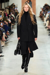 Model walks the runway in a black double-breasted coat, black turtleneck, knee-high boots, and a black handbag.