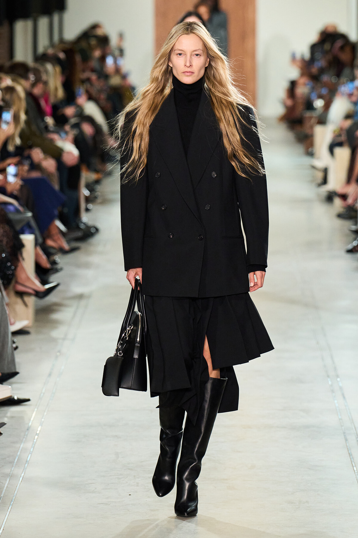 Model walks the runway in a black double-breasted coat, black turtleneck, knee-high boots, and a black handbag.