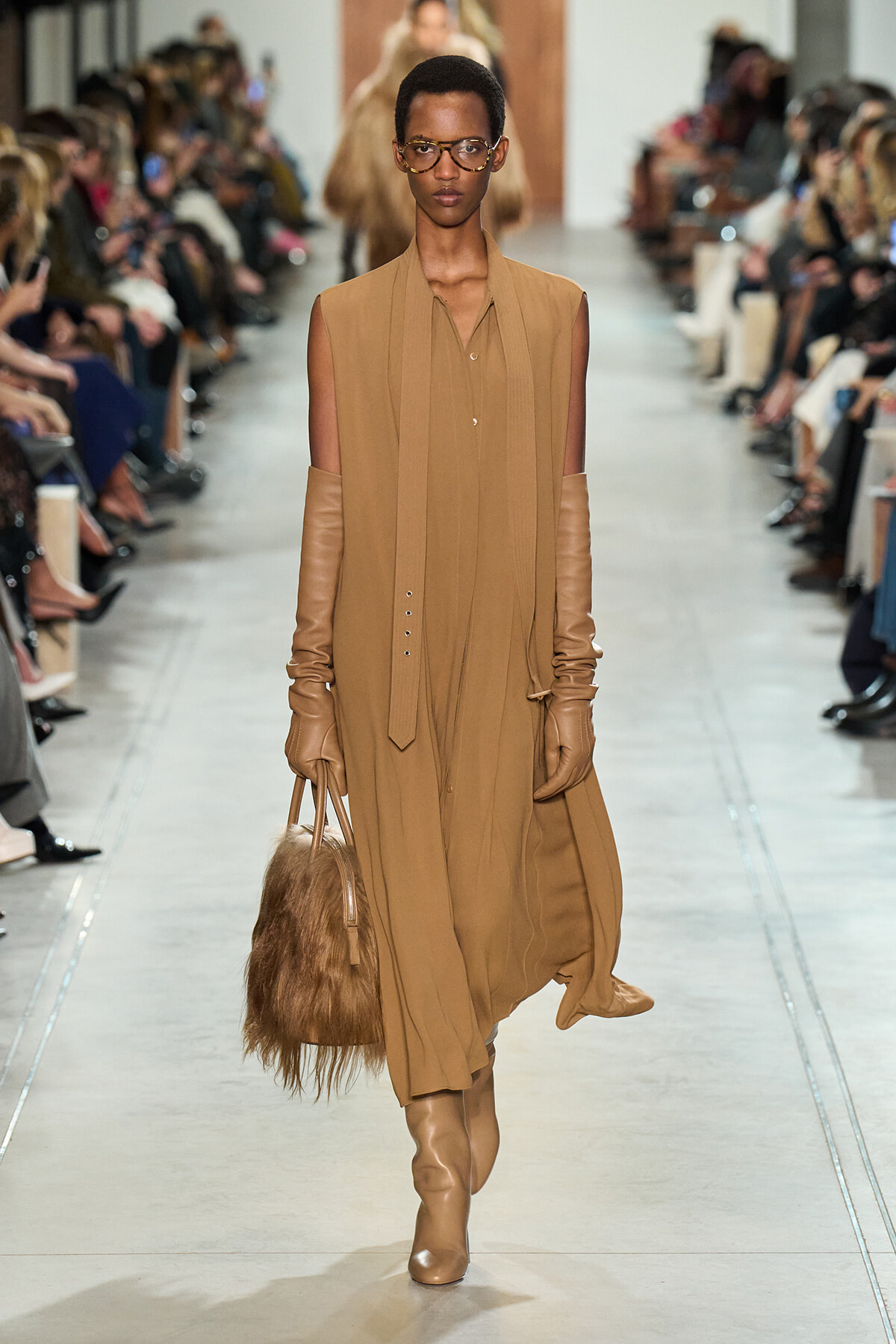 Model walks a runway in a camel-toned sleeveless dress with matching long gloves, tan boots, and a furry brown handbag.