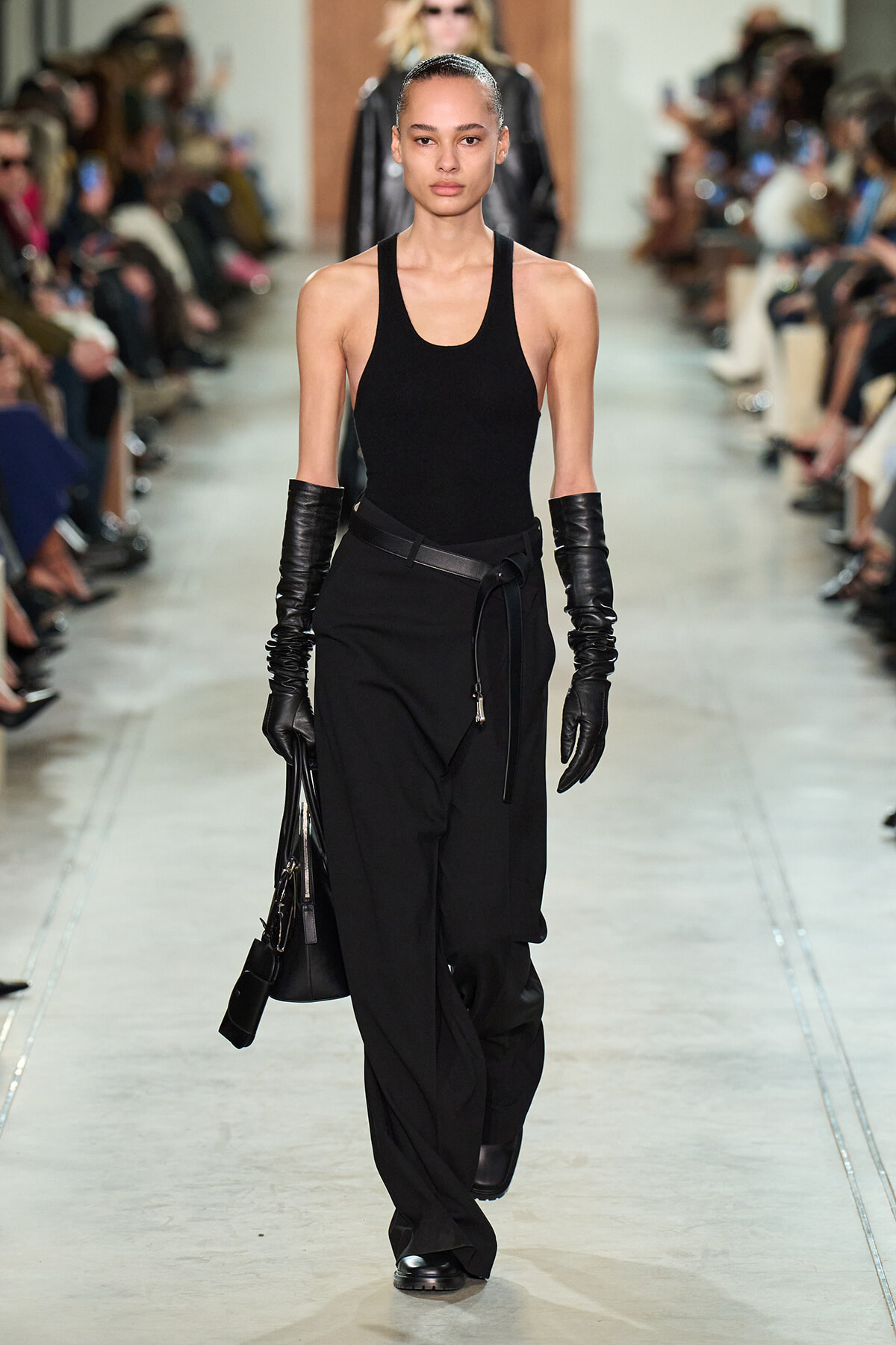 Model walking a fashion show runway in an all-black outfit: sleeveless top, wide-leg pants with belt, long leather gloves, and a black bag.