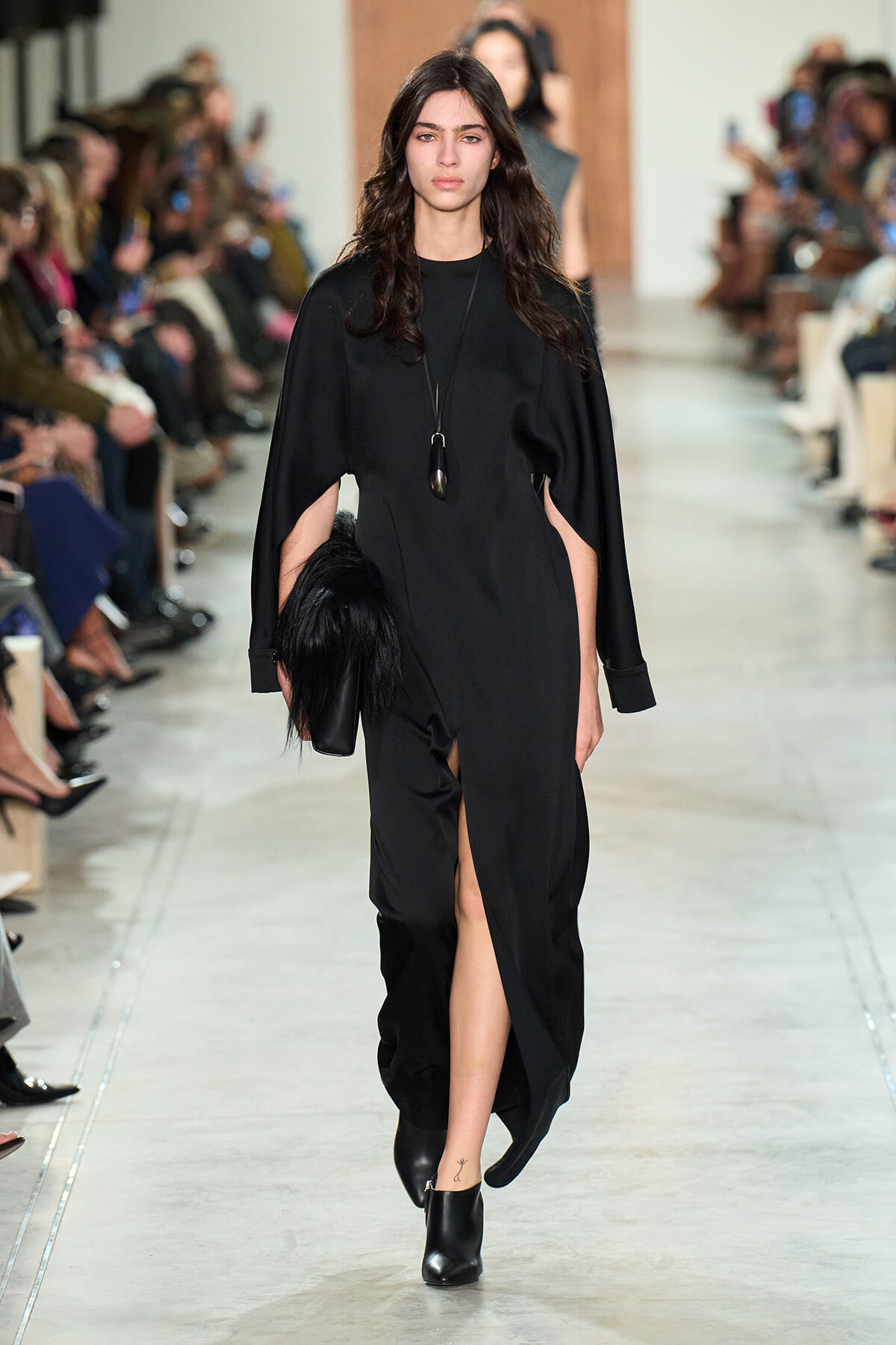 Model walks a runway in a long black dress with cape sleeves and a high slit, wearing a pendant necklace.