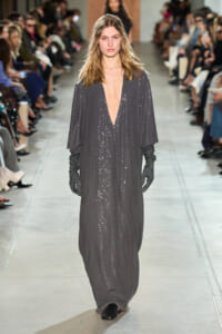 Model walks the runway in a dark gray, floor-length sequin gown with a deep V neckline and long black gloves.