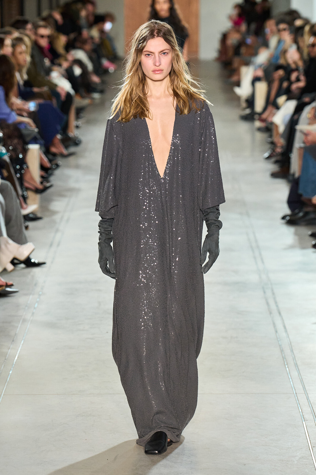 Model walks the runway in a dark gray, floor-length sequin gown with a deep V neckline and long black gloves.