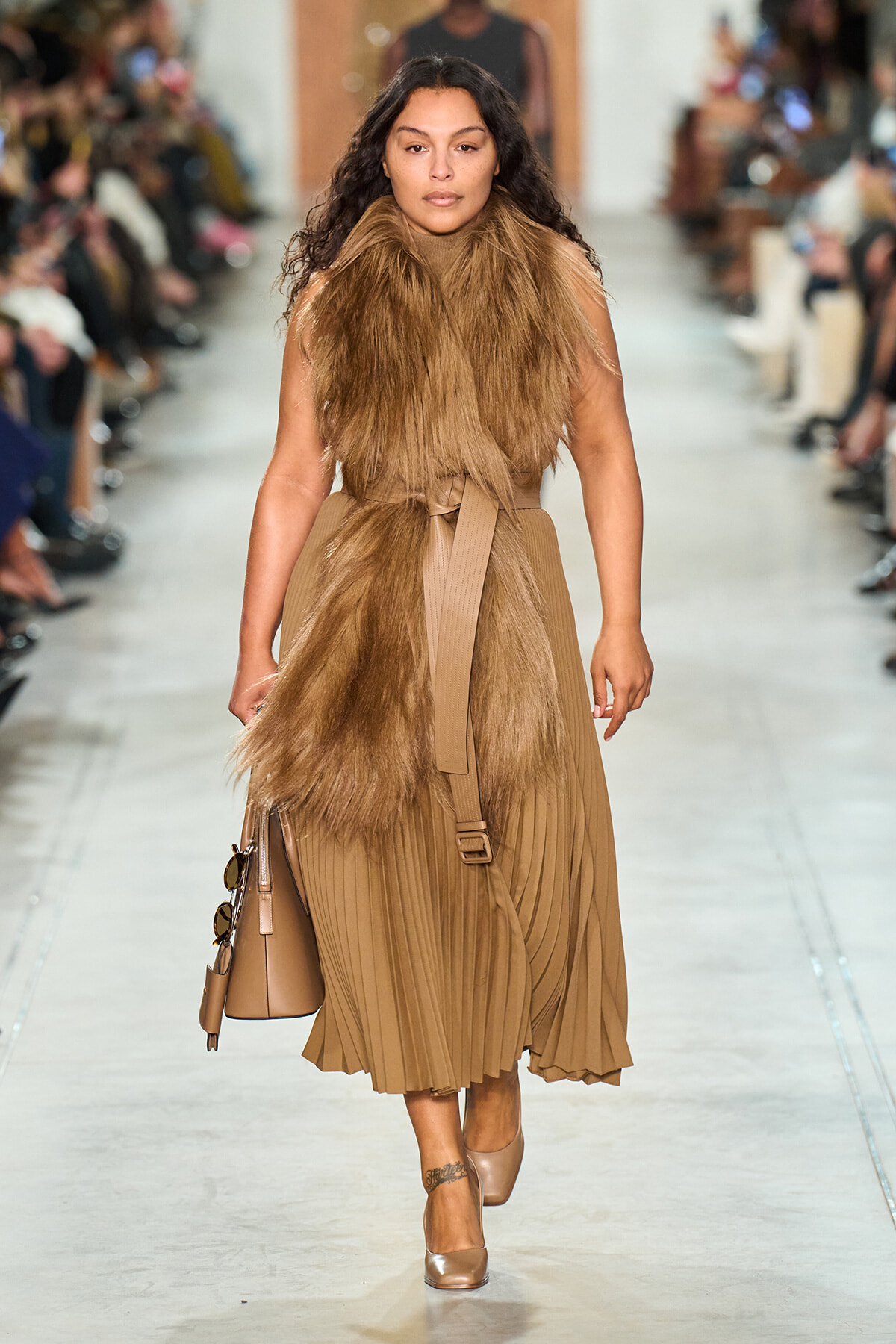 Model on a runway wearing a camel fur stole over a tan pleated midi dress, carrying a beige handbag.