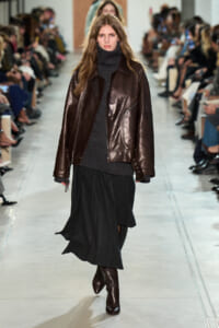 Model walks a fashion show wearing a dark brown leather jacket, black turtleneck, and a long black skirt.