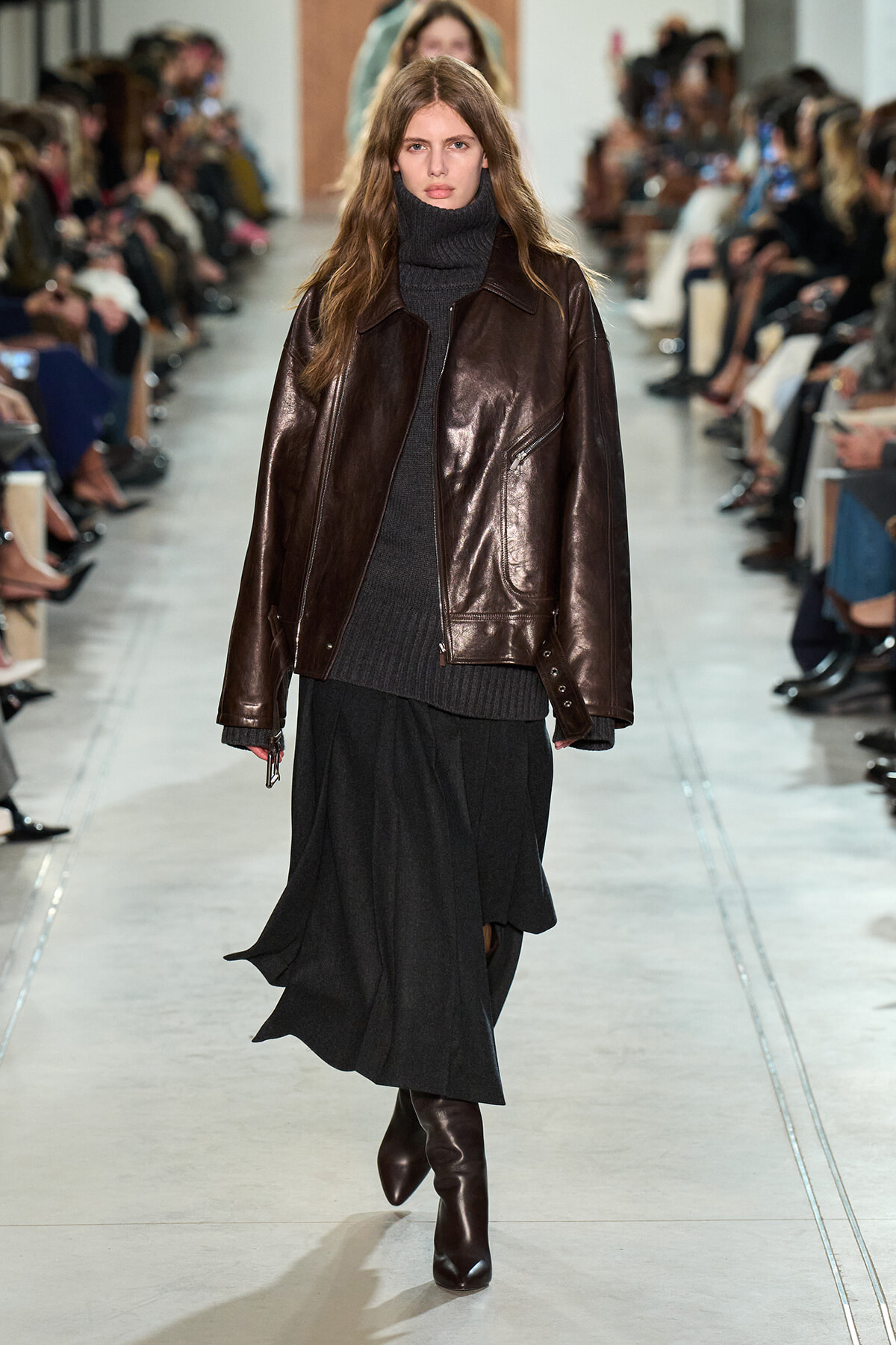 Model walks a fashion show wearing a dark brown leather jacket, black turtleneck, and a long black skirt.