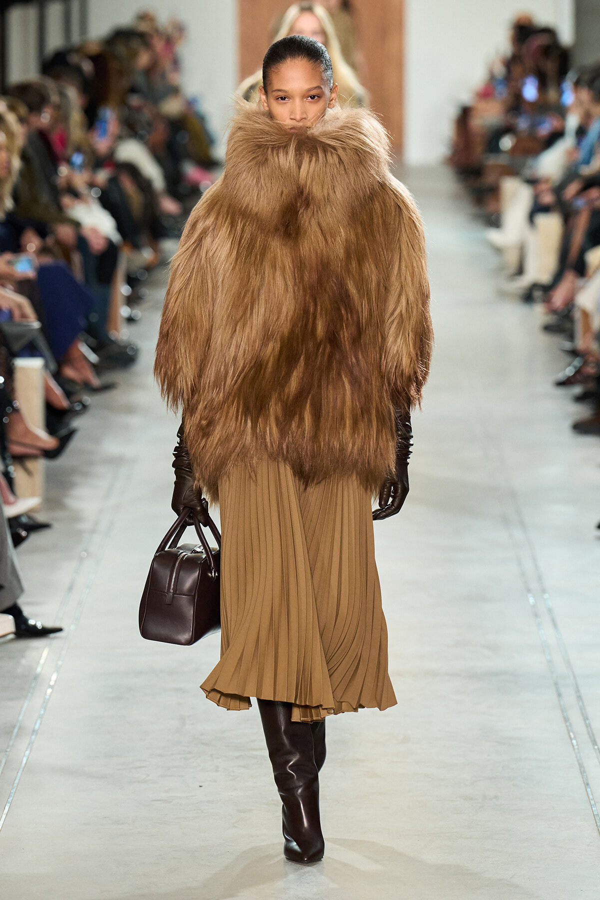 Model walks the runway in a voluminous brown fur coat over a tan pleated midi dress, black gloves, knee-high boots, and a dark brown handbag.