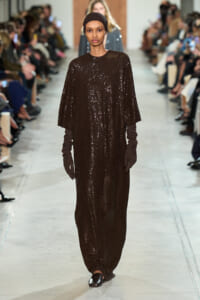 Model walks the runway in a full-length dark brown sequined dress with cape sleeves, matching gloves, and a black headscarf.