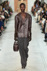 Model walks the runway in a brown crocodile-textured top, long brown gloves, gray wide-leg pants, and a brown crocodile bag with a pendant necklace.