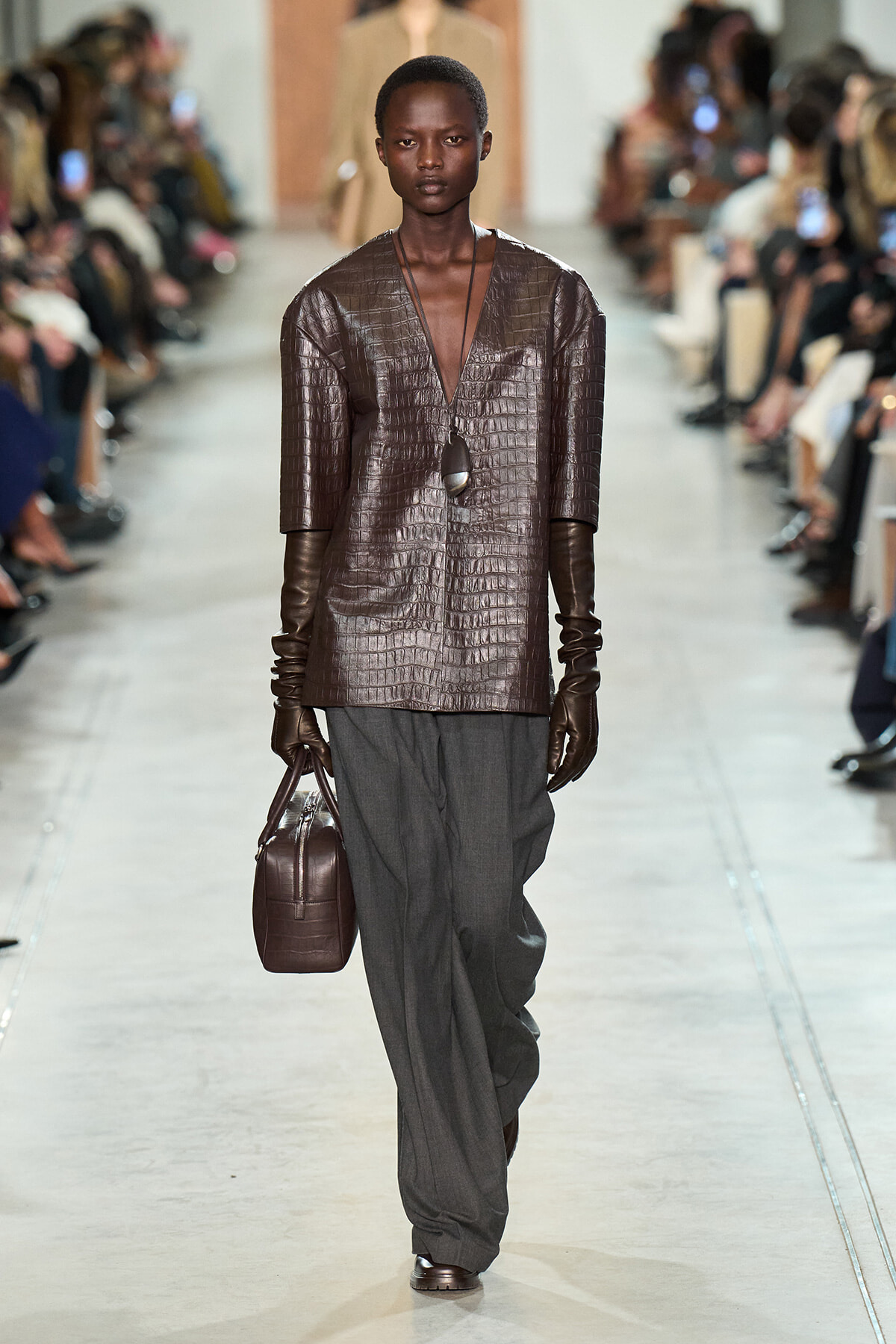 Model walks the runway in a brown crocodile-textured top, long brown gloves, gray wide-leg pants, and a brown crocodile bag with a pendant necklace.
