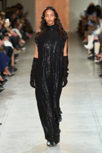 Model walks a runway in a floor-length black sequin gown with a high neckline and long black gloves, onlookers blurred in background.