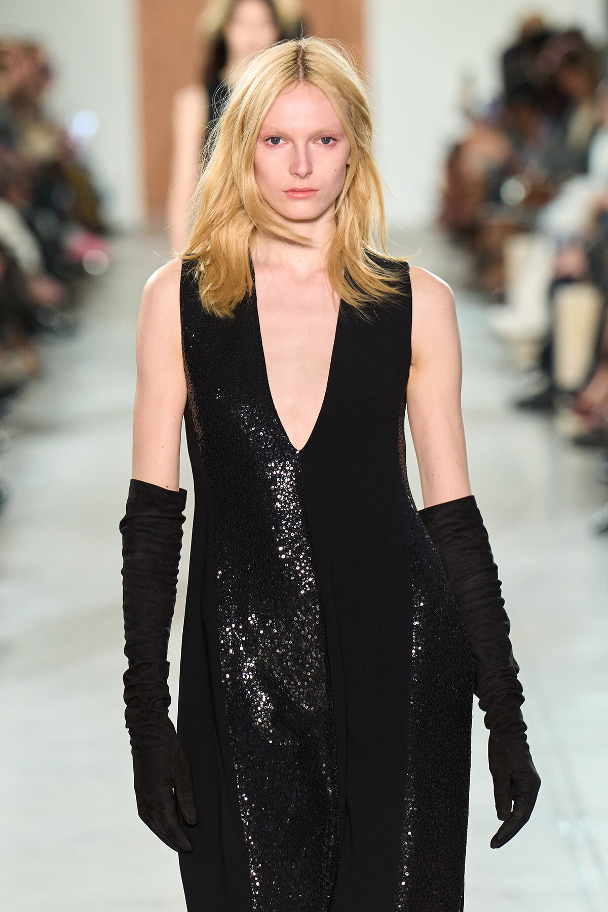 Blonde model walks the runway in a sleeveless black sequined dress with a deep V neckline and long black gloves.
