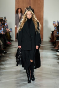 Model walking the runway in a black double-breasted coat, black turtleneck, and knee-high boots, carrying a black handbag.