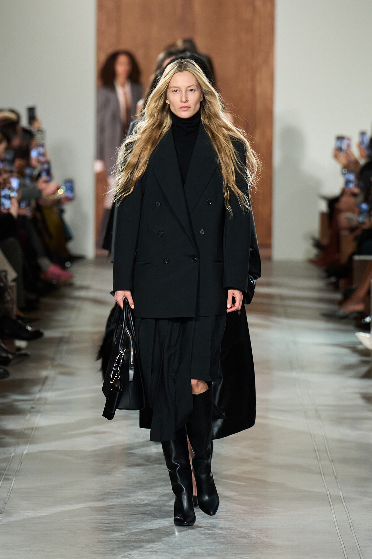 Model walking the runway in a black double-breasted coat, black turtleneck, and knee-high boots, carrying a black handbag.