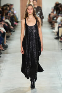 Model walks the runway in a long black sequined dress with black boots, audience blurred in the background.