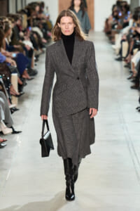 Model walking a runway in a grey plaid blazer and matching skirt with a black turtleneck and knee-high boots, carrying a black handbag.