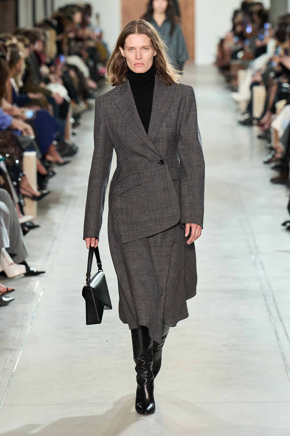 Model walking a runway in a grey plaid blazer and matching skirt with a black turtleneck and knee-high boots, carrying a black handbag.