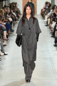 Model on a runway wearing a loose gray dress with wide sleeves, black gloves, a black clutch, and a long pendant necklace; audience sits along the catwalk.