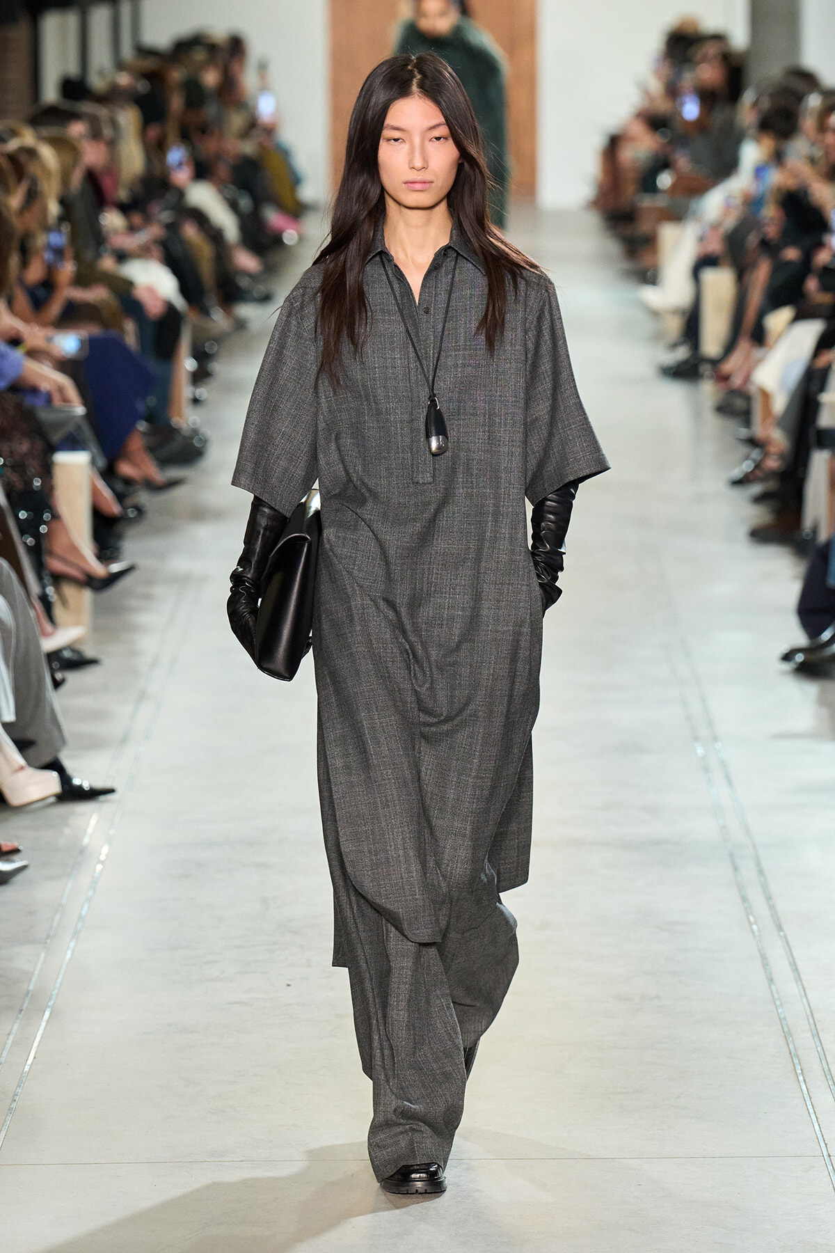Model on a runway wearing a loose gray dress with wide sleeves, black gloves, a black clutch, and a long pendant necklace; audience sits along the catwalk.