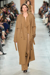 Model walks the runway in a long beige trench coat with layered lapels and belt.