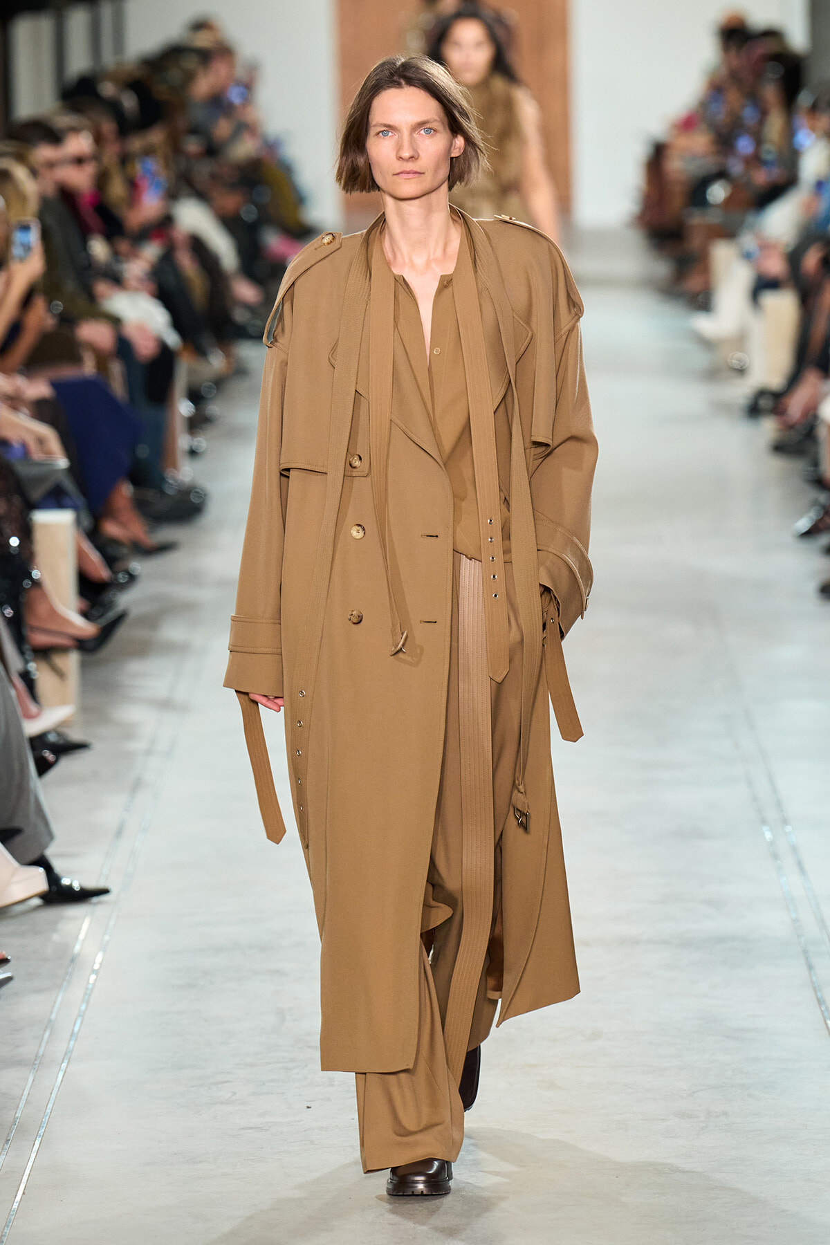 Model walks the runway in a long beige trench coat with layered lapels and belt.