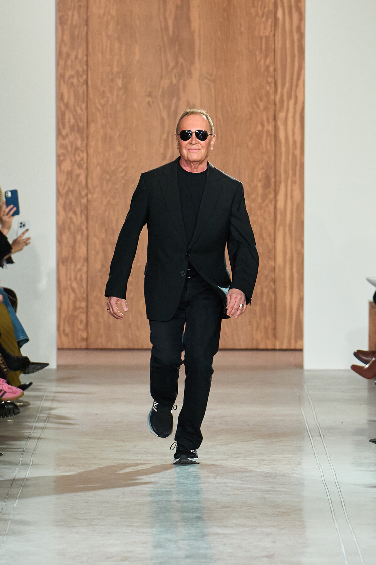 Man in a black suit and sunglasses walks down a fashion runway with a confident stride.