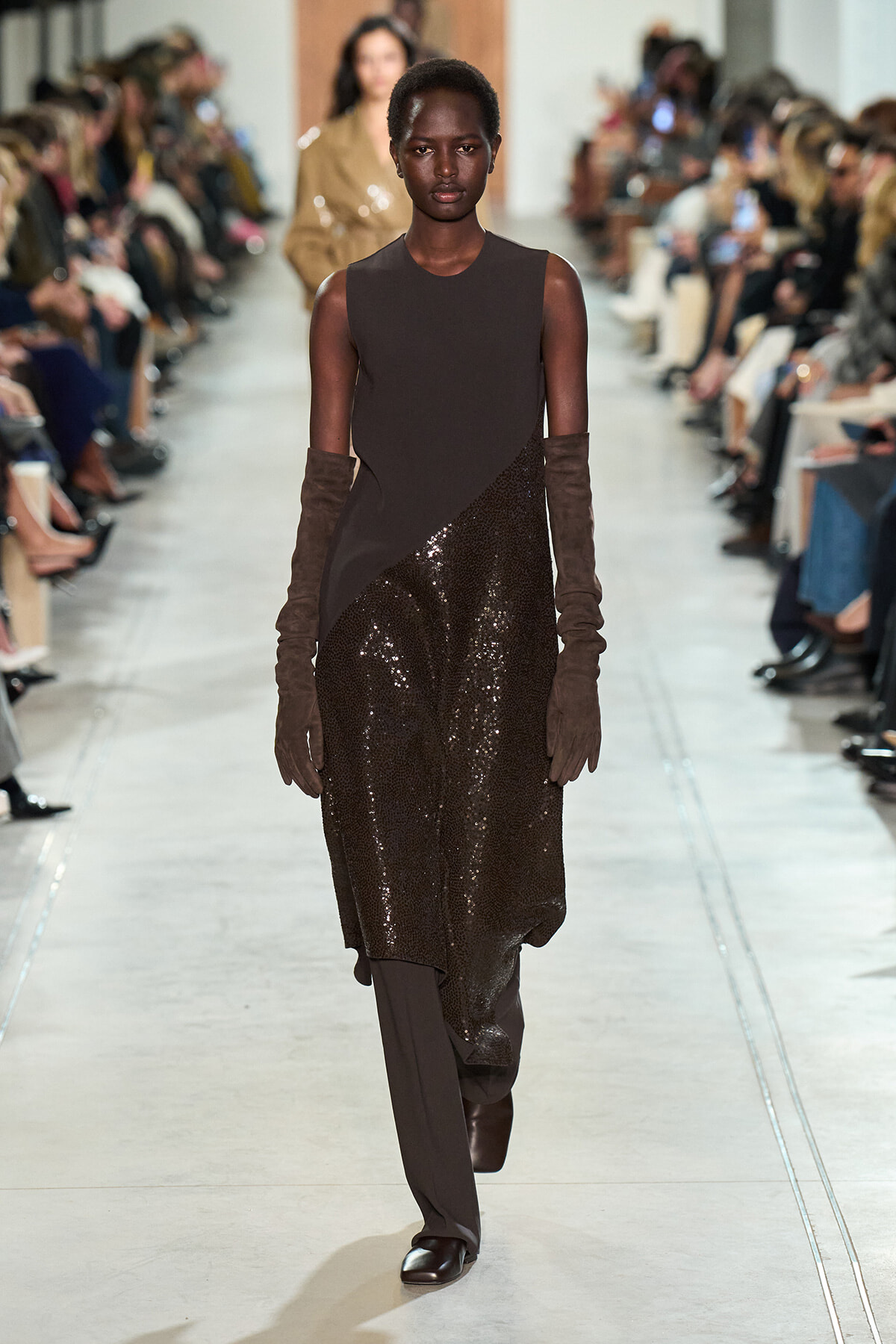 Model walks a fashion runway in a sleeveless dark brown top with a sequined brown skirt, paired with long brown gloves and boots.