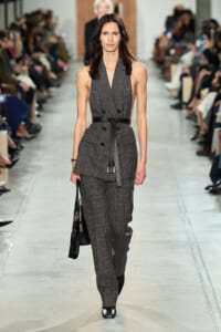 Model on a runway wearing a gray plaid sleeveless blazer with a black belt and matching pants, carrying a black handbag.