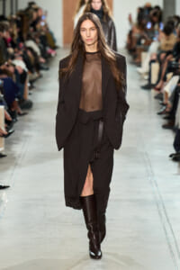 Model walks a fashion runway in a sheer black top, oversized blazer, and a black belted skirt with tall dark boots.