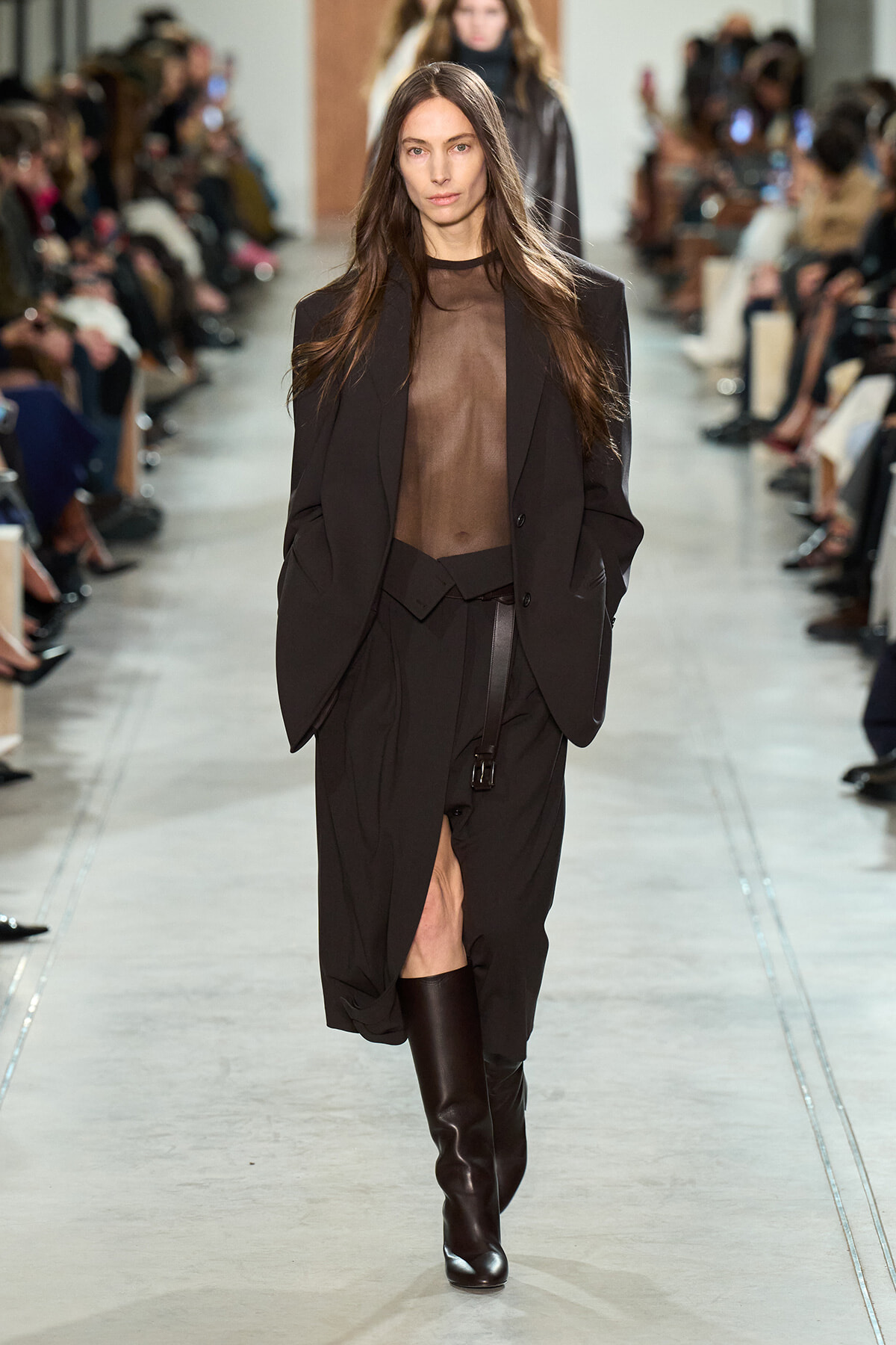 Model walks a fashion runway in a sheer black top, oversized blazer, and a black belted skirt with tall dark boots.
