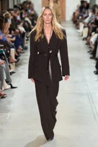 Model walks the runway wearing a black belted blazer and matching wide-leg trousers on the catwalk.